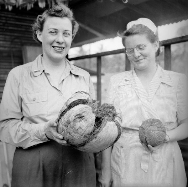 Lieutenant Jeanette Loree of East Lansing, Michigan (left) and Lieutenant Dorothy Watts of Jackson, Michigan (right), pose with a coconut and its husk. A building is behind them.