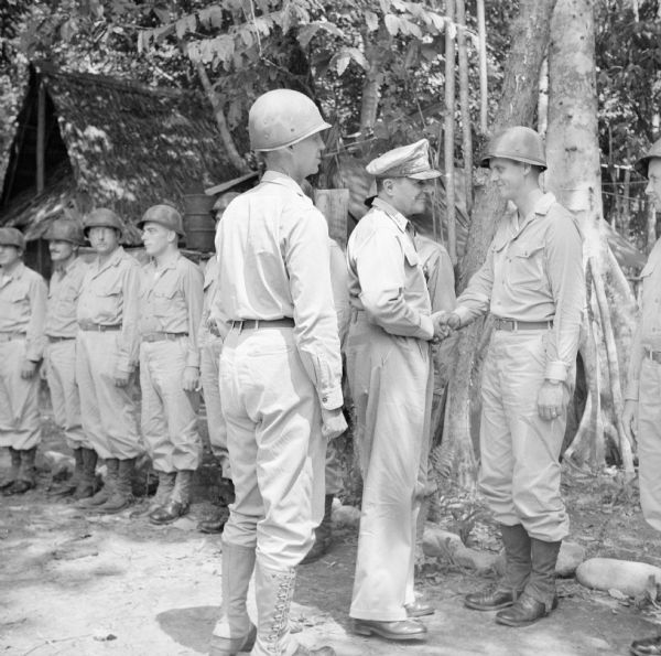 Four Star General Douglas MacArthur greets a soldier during a tour on Goodenough Island, in the Solomon Sea, New Guinea (present day Papua New Guinea). Other soldiers stand at attention in the background.