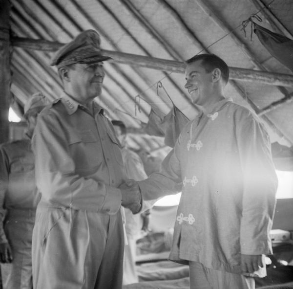 Four Star General Douglas MacArthur greets Lieutenant Lyle Hershey of Lansing, Michigan, during a tour on Goodenough Island, in the Solomon Sea, New Guinea (present day Papua New Guinea). They appear to be in a hospital setting. The soldier is wearing patient pajamas and beds are in the background.