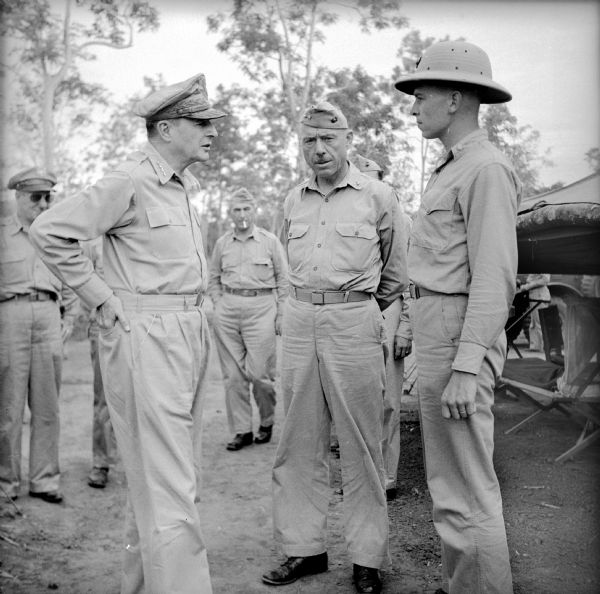 Four Star General Douglas MacArthur greets a soldier during a tour on Goodenough Island, in the Solomon Sea, New Guinea (present day Papua New Guinea). Soldiers and officers are standing in the background, and a tent is on the right.