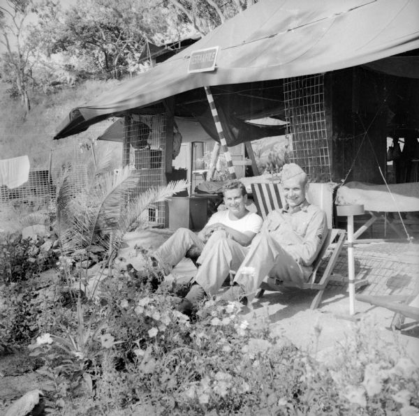 Technical Sergeant Leonard Stoltz (left) and Technical Sergeant Warren Fuhrman (right), both of Milwaukee, Wisconsin, relax in front of a tent at a base in New Guinea (present day Papua New Guinea). A sign that reads "Rest-A-While" hangs on the tent over their heads. There are plants and flowers in a garden at their feet.