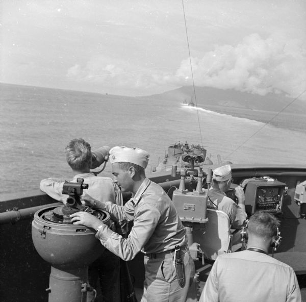 Navigation instruments on an American cruiser were checked by Lieutenant Commander Leonard Meyer of Appleton. Three other crew members are nearby. The cruiser took part in the heavy bombardment of Cape Gloucester, New Britain, New Guinea (present day Papua New Guinea), before the marines landed. In the distance are two ships, and beyond is smoke from the bombardment near the shoreline.