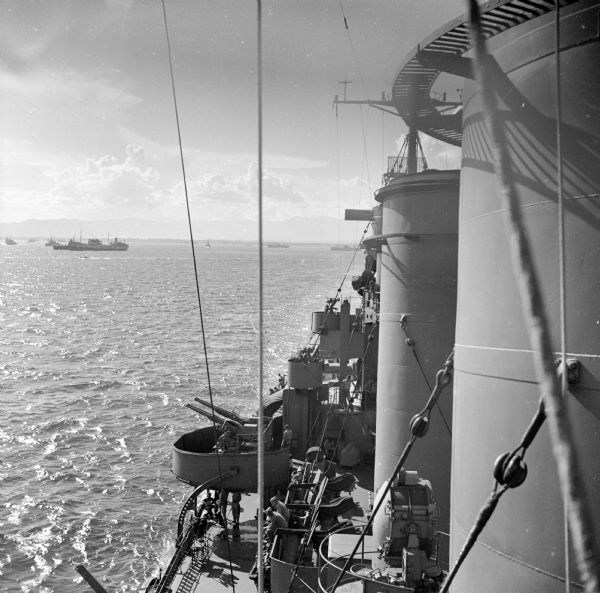 Elevated view looking down towards deck of an American cruiser leaving the base heading for the bombardment of Cape Gloucester, New Britain, New Guinea (present day Papua New Guinea), before the marines landed. In the distance are several warships, with land just visible in the distance.