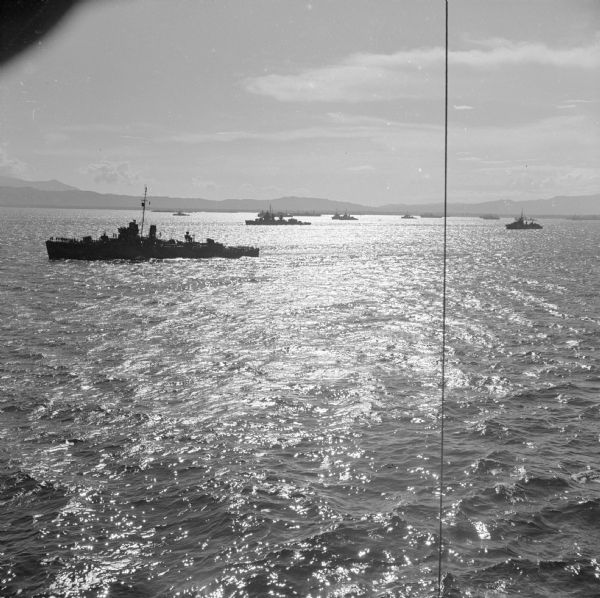 Elevated view, perhaps from an American Cruiser, of warships leaving the base heading for the bombardment of Cape Gloucester, New Britain, New Guinea (present day Papua New Guinea), before the marines landed. In the background is a mountainous shoreline.