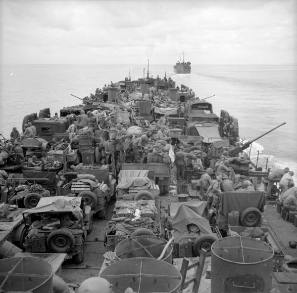 Elevated view of the loaded deck of a LST (Landing Ship, Tank) on the way from Goodenough Island to Saidor, New Guinea (present day Papua New Guinea). On the deck are many soldiers, jeeps, trucks, artillery and supplies. Tanks were probably in the hold of the ship. Another warship is in the distance. Robert Doyle wrote a caption for this image although it was not published at that time: "Packed with vehicles and soldiers, landing ship heads north for assault on Saidor, New Guinea, by task force consisting mainly of elements of Thirty-second Division. Landing was made at Saidor Jan. 2."