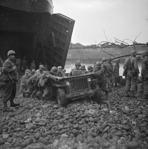 Soldiers push a jeep that is stuck on the rocky beach at the Saidor landing. The open front of the LST (Landing Ship, Tank) is on the left. Another LST can be seen on the right. Robert Doyle wrote a caption for this image although it was not published at that time: "When jeeps pulling loaded trailers dug into rocks and stalled, soldiers quickly pushed them out to clear the way for vehicles behind."