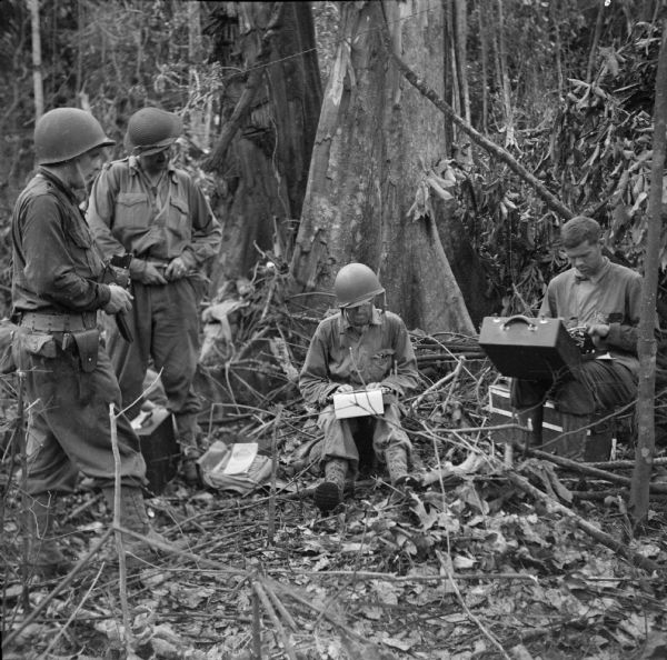 Three war correspondents at Saidor, New Guinea (present day Papua New Guinea). Names (left to right) are, unknown man with camera, John Scarlett of Australia, Ralph Teatsorth of United Press and Arthur Vesey of the <i>Chicago Tribune</i>. Three of the men are wearing combat gear with helmets. Two men have their typewriters perched on their knees. The jungle is all around them. Doyle's caption that ran with the photo: "Not much like the news room at the Journal, is it? Newsmen are pounding out stories two hours after the Red Arrow landing at Saidor."