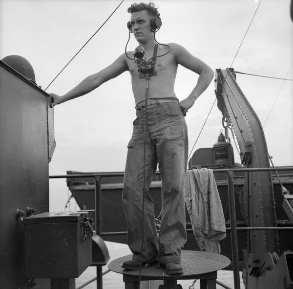 Seaman First Class Thomas Veater (shirtless) of Louzerne, Pennsylvania, on a LST (Landing Ship, Tank) after the landing at Saidor, New Guinea (present day Papua New Guinea). He is wearing headphones and a microphone.