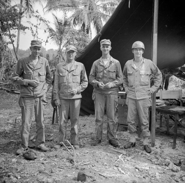 Four Army officers pose for a group portrait at a military camp on the north coast of New Guinea (present day Papua New Guinea). Names (left to right) are Major William Hastings of Corsicana, Texas, Lieutenant Colonel Tyler D. Barney of Sparta, Wisconsin, Captain Leonard Garrett and Major Charles (Tex) Meyer, both of Beaumont, Texas. A tent with table and chairs is on the right. Trees are in the background.