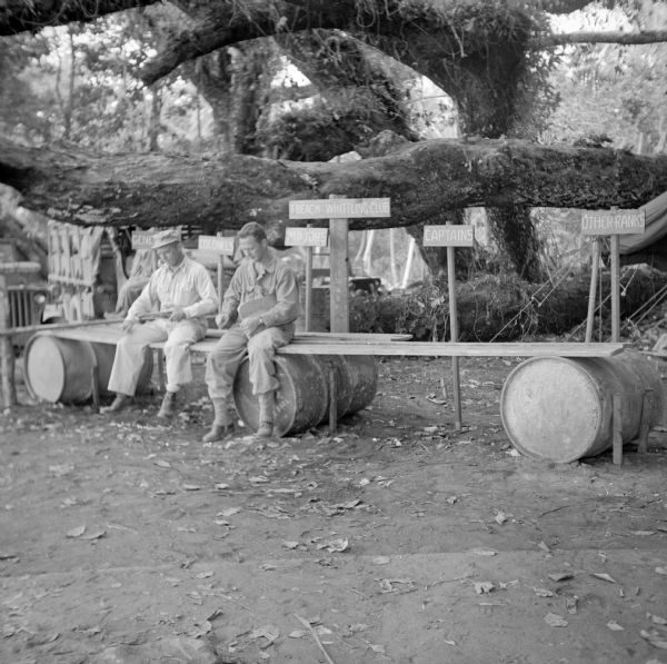 Two officers sit on improvised seats (boards on barrels) at the T-Beach Whittling Club, located on the north coast of New Guinea (present day Papua New Guinea). On the left is Lieutenant Colonel Hugh Fanning of LaCrosse, Wisconsin and on the right is Major Meredith Huggins of Salem Oregon. Major Huggins was awarded the Distinguished Service Cross, Silver Star and a Purple Heart. Signs behind them indicate where the different ranks should sit. The signs read, "Generals," "Colonels," "Majors," Captains" and "Other Ranks." Large trees with thick limbs are in the background. On the left are jeeps and on the right is a tent.