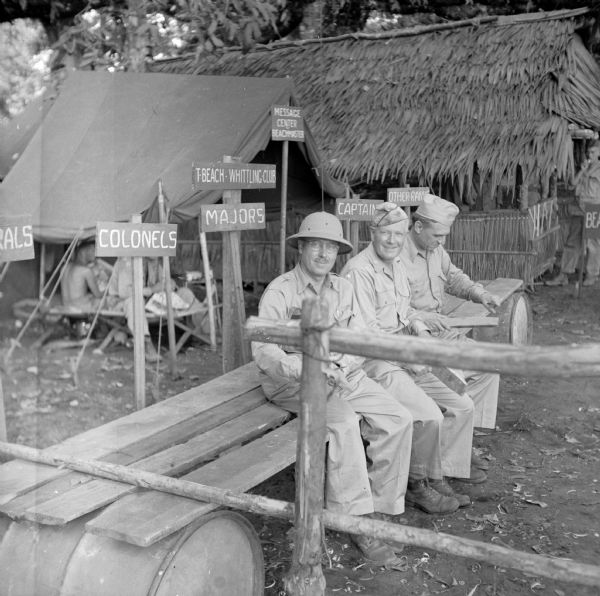 Three men sit on improvised seats (boards on barrels) at the T-Beach Whittling Club, located on the north coast of New Guinea (present day Papua New Guinea). They are (left to right) Frank E. Mason, Special Assistant to the Secretary of the Navy from Milwaukee, Wisconsin, Lieutenant Colonel Phillip F. La Follette of Madison, Wisconsin, and Raymond Clapper, a columnist. They were on their way from Cape Gloucester to Milne Bay and Guadalcanal in the Solomon Sea. Signs behind them indicate where the different ranks should sit. The signs read, "Generals," "Colonels," "Majors," Captains" and "Other Ranks." A tent, with soldiers sitting on cots, with a sign "Message Center Beachmaster," is behind them and on the right is a building built in the indigenous style. Special note by Robert Doyle: "Frank E. Mason is a Vice President of NBC (on leave) and is the son of the late Dr. George Franklin Mason." Lieutenant Colonel Phillip F. La Follette was three times Governor of Wisconsin.