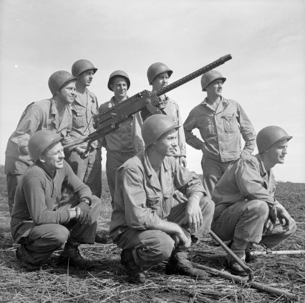 Eight soldiers from Wisconsin pose with a captured Japanese gun. It may be a Japanese 12.7-mm (Fixed Mount) Aircraft Machine Gun. The soldiers were stationed at the military camp at Saidor, New Guinea (present day Papua New Guinea). Their names (kneeling, left to right) are Private First Class Eugene Kobza of Milwaukee, Sergeant LaVerne Ader of Almond and Corporal John Malecki of Berlin; (standing, left to right) are Sergeant Delbert DeForest of Fort Atkinson, Corporal Eugene Weinand of Jackson, Sergeant Charles Allhands of Madison, Private Leonard Bieganowski of Milwaukee and Sergeant Ward K Wentworth of Whitewater.