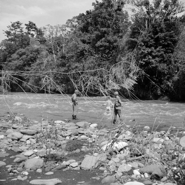 Three soldiers view an old damaged indigenous suspension bridge. The soldier with a rifle on the right is Private Marion Tewalt of Viroqua, Wisconsin. The naked soldier wading in the water is Private Jasper Edwards of Durham, North Carolina. The standing soldier on the left is Private Robert Anderson of Chippewa Falls, Wisconsin. The shore is rocky in the foreground and trees are on the far side of the river.