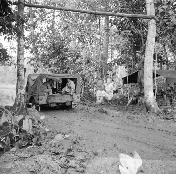 Sergeant Walter Schaefer of Port Washington, Wisconsin, swings from a rope over an extremely muddy road just after a truck has just driven through at a military camp at Saidor, New Guinea (present day Papua New Guinea). At least four soldiers are riding in the truck. Waiting on the pole for his turn is Sergeant Armond Lackas of Hartford, Wisconsin. A tent is on the right.