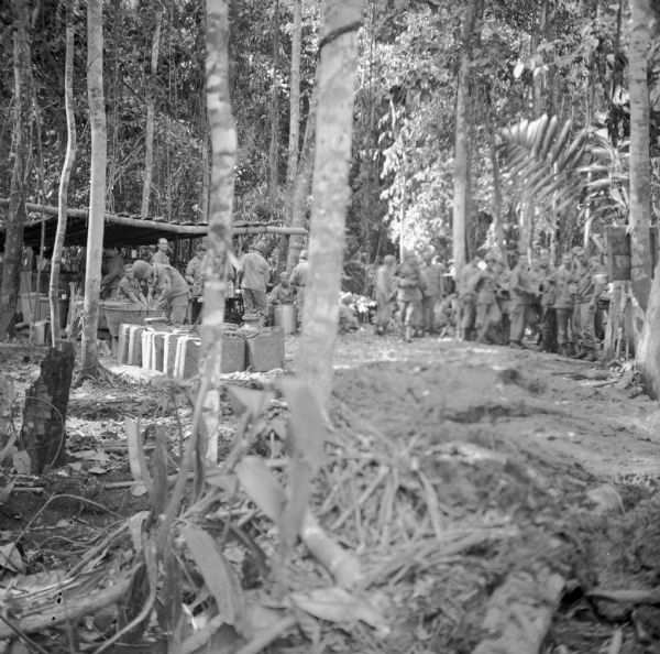 An outdoor kitchen scene at the military camp at Saidor, New Guinea (present day Papua New Guinea). Soldiers from the 121st Field Artillery Battalion are lined up waiting for the kitchen to announce the meal is ready. The kitchen is located under a roof with open sides, and water cans and large cooking vessels are nearby.