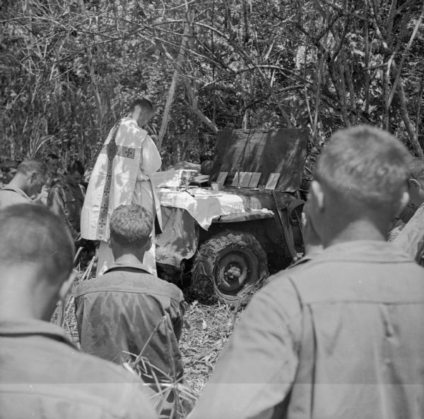 Catholic Mass at Saidor, New Guinea (present day Papua New Guinea). The Chaplain is using the hood of a jeep as an altar. Several soldiers can be are in the foreground worshipping. Trees are in the background. Robert Doyle wrote a caption for this image although it was not published at that time: "Army Jeep serves as altar at Catholic mass conducted by Father Anthony E. Burakowski, 30, Fairfield, Conn., Jan. 23 at artillery headquarters area at Saidor, New Guinea. Lieutenant Burakowski arrived at area in 'grasshopper' observation plane. It was his first plane ride. He arrived in New Guinea from America in December, 1943."