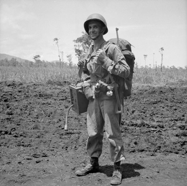 Private First Class Carl Krejci of Chippewa Falls, Wisconsin, carries a heavy load as he leaves on patrol from camp in Saidor, New Guinea (present day Papua New Guinea), for a week. In addition to his own gear and weapon, he appears to have a radio on a strap over his shoulder. A muddy, rutted road is behind him.