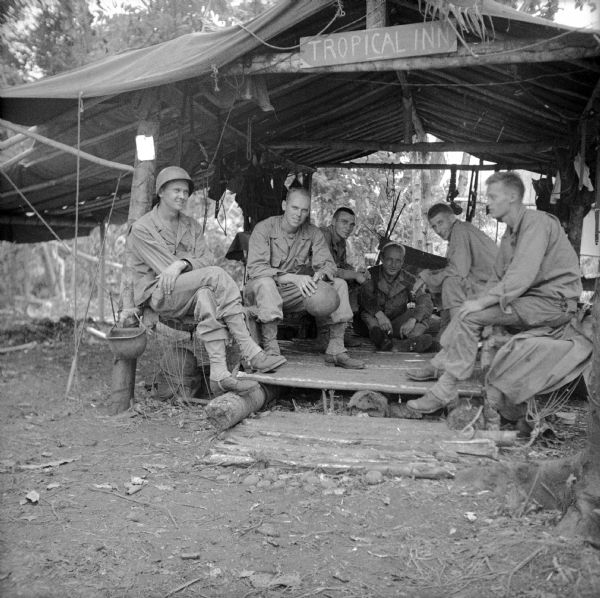 Soldiers and officers relax in the "Tropical Inn," a tent that has seating and a raised wooden floor, at Saidor, New Guinea (present day Papua New Guinea). The soldiers are, (left to right) Lieutenant Otto Shild of Gooding, Idaho, Sergeant Bill Howard of La Crosse, Wisconsin, Sergeant Edwin Morse of La Crosse, Wisconsin, Corporal Edward Kukkola of Kelso, Washington, Corporal Frank Schroeder of La Crosse, Wisconsin and Private First Class Dorrison Buros of Viroqua, Wisconsin.