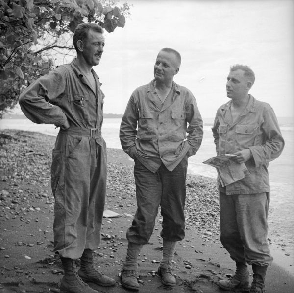 Wisconsin artillery officers helping direct the shelling of the Japanese on Saidor's west flank (beyond the point in the background). The officers are, (left to right) Major Robert Schultz of Milwaukee, Lieutenant Colonel Erwin Beyer of Madison and Major Francis Knope of Stevens Point. A tree is on the left, beach and ocean in the background. Saidor is located on the northeast coast of New Guinea (present day Papua New Guinea).