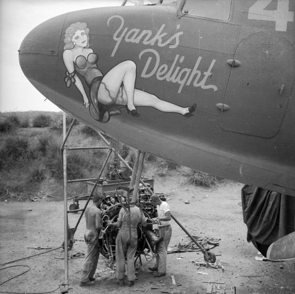 Elevated view of three soldiers working on the engine of a warplane at an airbase near Port Moresby, New Guinea (present day Papua New Guinea). The plane has nose art depicting a reclining woman in the "pin-up style" with the text, "Yank's Delight."