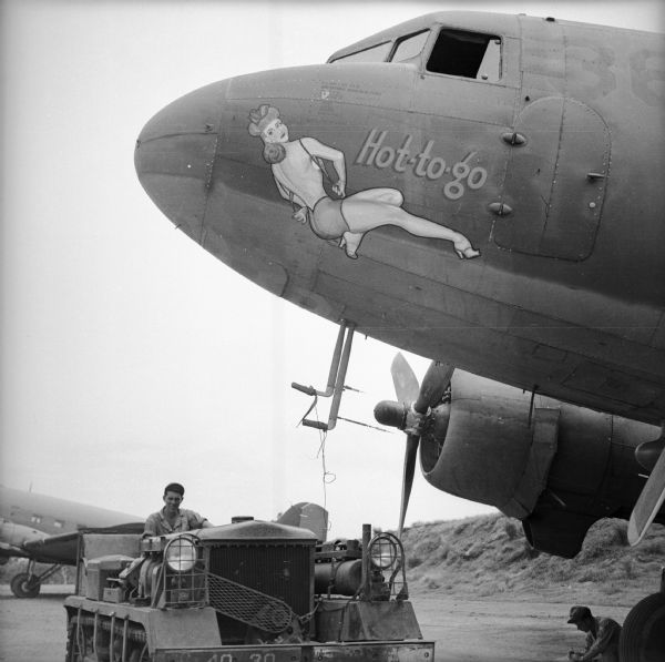 A soldier sits in a bulldozer parked under the nose of a warplane at an airbase near Port Moresby, New Guinea (present day Papua New Guinea). Another man is crouched near the wheel of the airplane on the right. The warplane has nose art depicting a reclining woman in the "pin-up style" with the text, "Hot-to-go." There is another airplane in the background.