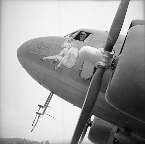 A pilot peers out of the window of a warplane at an airbase near Port Moresby, New Guinea (present day Papua New Guinea). The plane has nose art depicting a reclining woman in the "pin-up style" with the text, "Open Date."