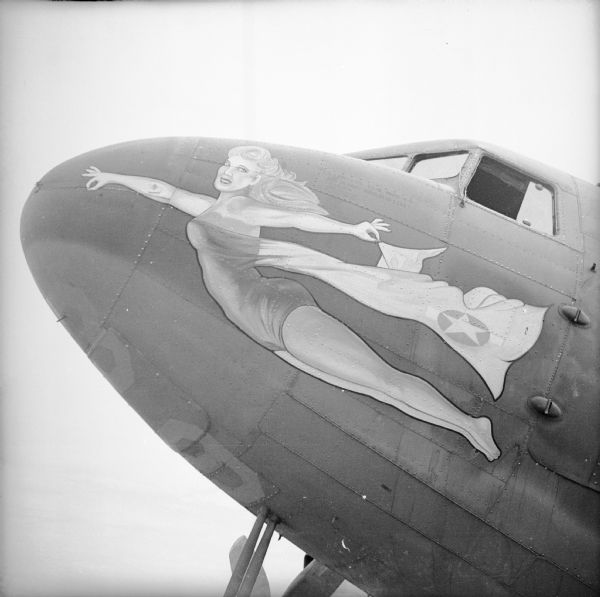 A warplane with nose art depicting a flying woman in the "pin-up style," at an airbase near Port Moresby, New Guinea (present day Papua New Guinea). She is wearing a strapless, one-piece swimsuit, and a cape with the Army Air Force star on it.