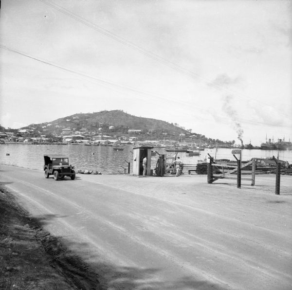 General view of Port Moresby Harbor from road, New Guinea (present day Papua New Guinea). A jeep is on the road on the left. Several soldiers are near a shack at the gate opening onto the Royal Australian Air Force Marine Section Parking Area. On the far right, several warships are docked. Across the harbor industrial buildings are along the shoreline at the base of a hill.