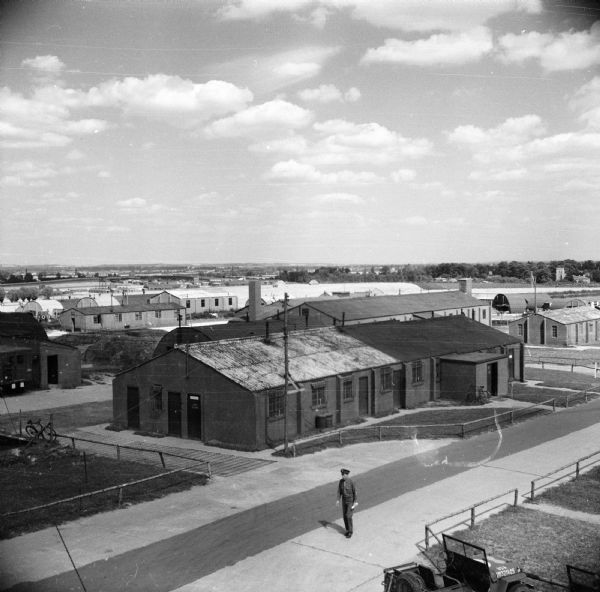 Elevated view of the buildings at the airfield at Royal Air Force station Steeple Morden, located 3.5 miles west of Royston, Hertfordshire, England. The 355th Fighter Group was stationed there. Bicycles are parked near buildings. Major Daniel M. Lewis of Ladysmith, Wisconsin, is walking on the road in the foreground. A jeep is parked in the lower right corner.