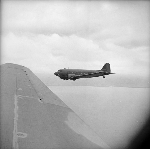 Aerial view over wing of formation of Douglas C-47 Skytrain cargo and troop transport planes leaves Saidor, New Guinea (present day Papua New Guinea). Robert Doyle notes that they were off the Japanese-held coast. The wing of the plane in which Doyle was a passenger is in the foreground. The plane in the center has the nose art depicting a reclining woman in the "pin-up style" with the text, "Hot-to-go."