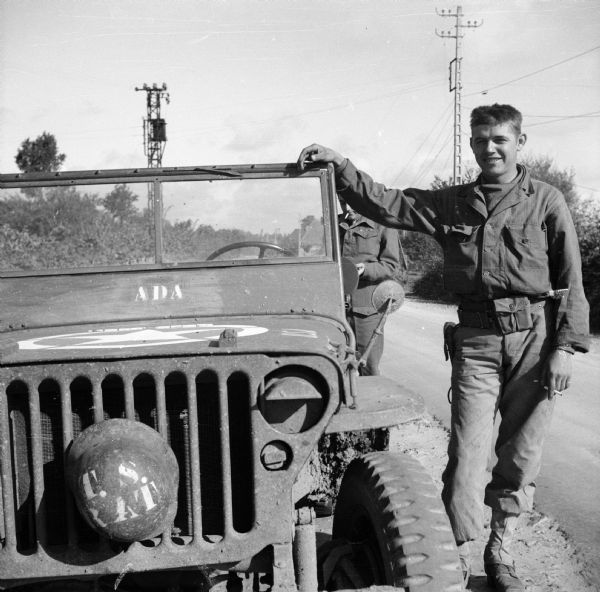 A roadside stop in Normandy, France. Two men are posing next to the parked jeep, which has "ADA" stenciled below the windshield. The man in front partially obscures the other man standing behind him. One man is Eric Baume, Australian correspondent and London Bureau Chief for <i>Truth Newspapers</i>. The other is Edward Chichester, the 6th Marquess of Donegall, ace reporter for the <i>London Sunday Dispatch</i>. A helmet is attached to the grill of the jeep with the name, "T.S. Rat" stenciled on it. Power poles and lines are in the background.