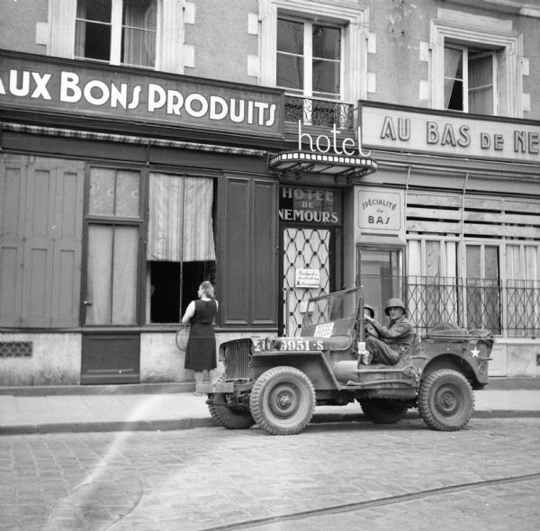 View across cobblestone street of a military jeep (marked "PRESS") is parked in front of the Hotel de Nemours, Rennes, France. Driving the jeep is Corporal Sidney Levine of Brooklyn, New York and the passenger is Private Fred Davis, Jr. of Pine Bluff, Arkansas. A woman on the sidewalk is standing at an open window behind them. Robert Doyle notes that the hotel had been "taken over by PRO (possibly Public Relations Office) for correspondents." This is where he stayed while in Rennes.