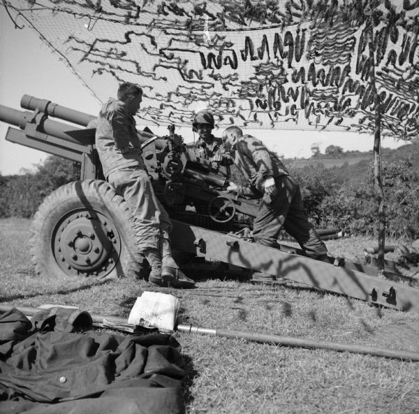 Soldiers with a 105mm Howitzer near Brest, France. Camouflage netting is over their heads. Names, (left to right) are Private First Class Thomas Greatorex of East Corinth, Maine, Sergeant Nick Falcone of Bangor, Pennsylvania and Private Emery Seidel of Milwaukee, Wisconsin. Hills and trees are in the background. Robert Doyle wrote a caption for this image although it was not published at that time, "Jolt for Jerry is served up by Private Emery Seidel, 31, (address), shown putting shell in breech of 105 millimeter howitzer outside Brest, France. Others in gun crew are (see above)."