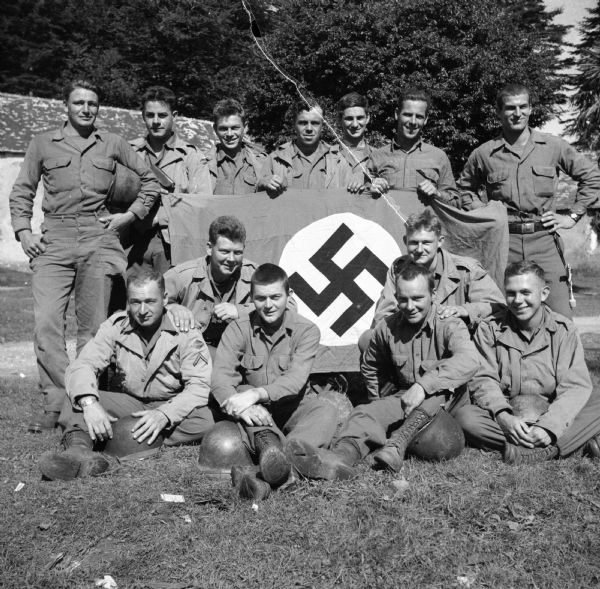 Robert Doyle wrote a caption for this image although it was not published at that time, "Rough and tough Wisconsin rangers, who helped storm Normandy beaches on D-Day and who had important assignment in capture of Brest, pose with Nazi flag and other souvenirs. Seated and kneeling are, (from left) Private First Class Oscar Behrent, 25, Elcho; Private Gerald Sejba, 25, Antigo; Private First Class Jack Thomas, 19, (address), Milwaukee; Private First Class Sigurd Sundby, 23, (address), Milwaukee; Private First Class Alvin Potratz, 19, (address), Milwaukee; and Private First Class Garness Colden, 20, Blanchardville. Standing are (same order) Private First Class George Frenchette, 20, Town of Greenfield; Private First Class David Neugent, 21, (address), Milwaukee; Private First Class Don Pechacek, 21, Ellsworth; Private First Class Jim Kohl, 27, River Falls; Private First Class Tom Armbruster, 19, Iron River; Private First Class John Tindell, 31, Platteville, and Sergeant Edward O'Connor, 21, Mosinee." A building and trees are in the background.