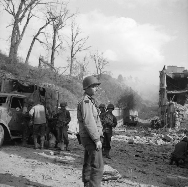 Robert Doyle views the destruction on a street in Brest, France. On the left is a damaged military truck with three soldiers standing. Behind Doyle are two more soldiers, one smoking a cigarette. On the far right a soldier is kneeling. In the background are the remains of a vehicle near a stone wall at the base of a hill.