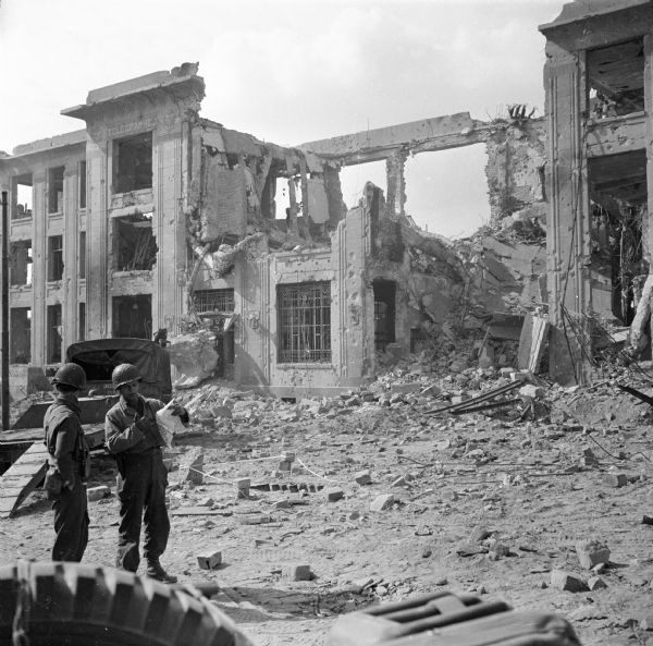 Two soldiers in the lower left look at a map. Behind them is what remains of the Brest Post Office. Just under the decorative stone cornice at the top is the word "TELEGRAPHES," indicating that the building also housed the telegraph office. Rubble fills the street. Behind the soldiers is an empty military flatbed truck. The tire and gas can in the extreme lower left may be the back of the jeep in which Robert Doyle was sitting when he snapped the photo.