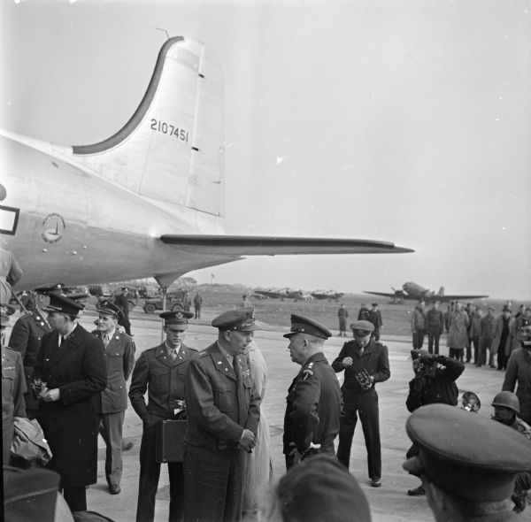 General Dwight D. Eisenhower speaks with General George C. Marshall at an airfield in Paris, France. On the left are more officers. Several photographers are on the right. Robert Doyle notes: "Arrival of first plane US ATC (United States Air Transport Command) to fly direct from America today." The airplane is a Douglas C-54 Skymaster. In the background are jeeps, more airplanes and a group of spectators.