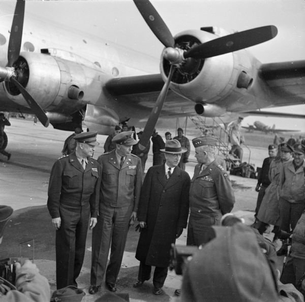 General Dwight D. Eisenhower (SHAEF commander) poses with (left to right) General George C. Marshall (Army Chief of Staff), Justice James F. Byrnes (Senior Advisor to President Roosevelt) and Lieutenant General Omar Nelson Bradley (Army Ground Commander) at an airfield in Paris, France. Several photographers can barely be seen in the foreground. A crowd watches from the right. Robert Doyle notes: "Arrival of first plane US ATC (United States Air Transport Command) to fly direct from America today." The airplane is a Douglas C-54 Skymaster.