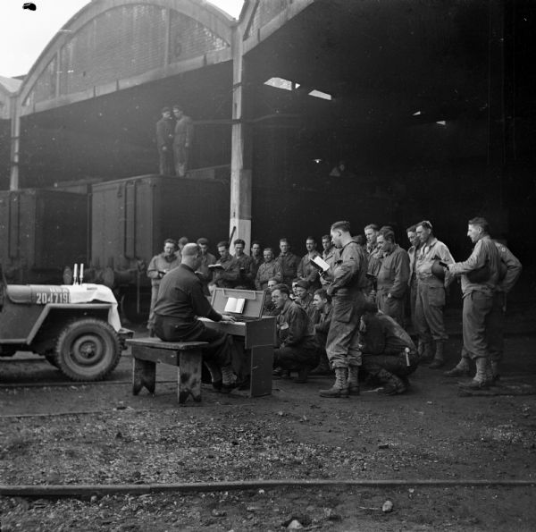 Chaplain Captain Allan R. Fredine holds a Protestant church service in a roundhouse used by the 757th Railway Shop Battalion in Cherbourg, France. He is using the hood of his jeep as an altar and is seated at a portable piano. Many soldiers stand and sit while singing from hymnbooks. In the upper left corner, two more soldiers look on from on top of a railroad car. Robert Doyle wrote a caption for this image although it was not published at that time, "Church services at the roundhouse are held for men of a railway shop battalion sponsored by the Milwaukee Road. Chaplain conducting this service near Cherbourg is Captain Allan Fredine of Minneapolis, Minnesota, who had Baptist churches in Eau Claire and Superior."