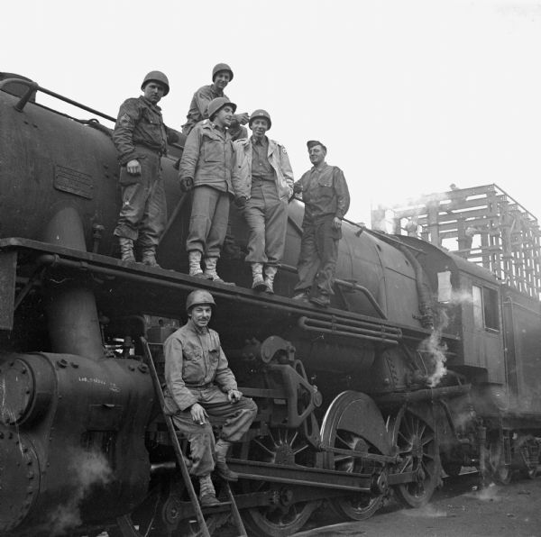 Five railroad soldiers pose on a locomotive near the roundhouse used by the 757th Railway Shop Battalion in Cherbourg, France. From Wisconsin are, (top) Sergeant Raymond Janiszewski of Milwaukee, (standing, left to right) Technical Sergeant Donald Fetzer of Manitowoc, First Lieutenant Sigmund Gralewicz of Milwaukee, Captain Earl D. Austin of Wauwatosa, and Sergeant Walter Demitros  of Milwaukee. Perched on the ladder is Staff Sergeant Robert Kurman, mechanic, of Brookyln, New York. A metal structure, with three soldiers working on it, is in the background.