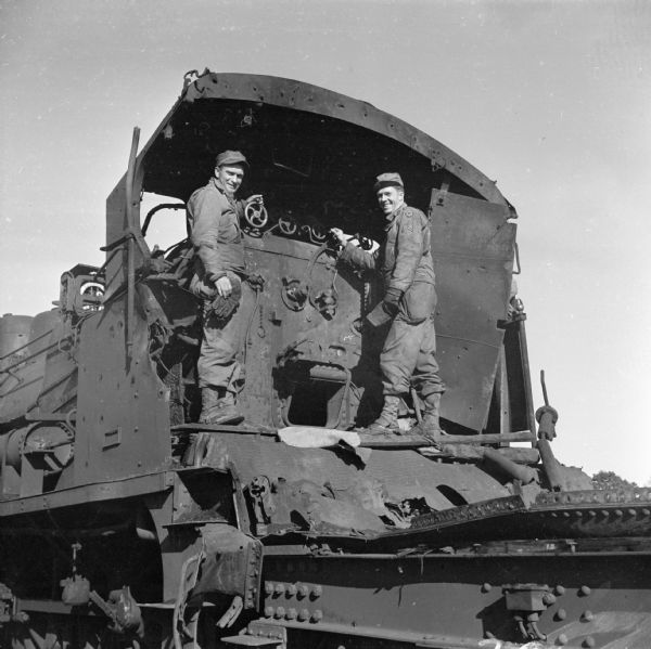 Two soldiers pose on a wrecked French locomotive in Folligny, France. Names, (left to right) are Switchman Private First Class Kenneth Walters of St. Paul, Minnesota and Yard Conductor Sergeant John Welch of Milwaukee, Wisconsin. Back home, Private First Class Walters was a switchman for the Northern Pacific. Robert Doyle wrote a caption for this image although it was not published at that time, "This may not be the 'Old 97', but the French locomotive is sufficiently wrecked to qualify it for the song. Damage was done at railroad yards in Normandy bombed by Allies before invasion. In the cab are (names above), members of railway operating battalion. Welch was a Milwaukee Road conductor before entering service."