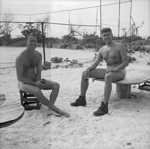 Two men enjoy a nude sun bath on Buster Island in the Kwajalein Atoll, Marshall Islands, in the South Pacific. The man on the right, wearing only boots, is Ensign Ray Hansen, NATS (Naval Air Transport Services) Administrative Officer of Milwaukee, Wisconsin. He is seated on a table, the other man is seated on a crate. Both of them have a beverage. The ground appears to be sand and a sports area is fenced off behind them with foliage in the background.