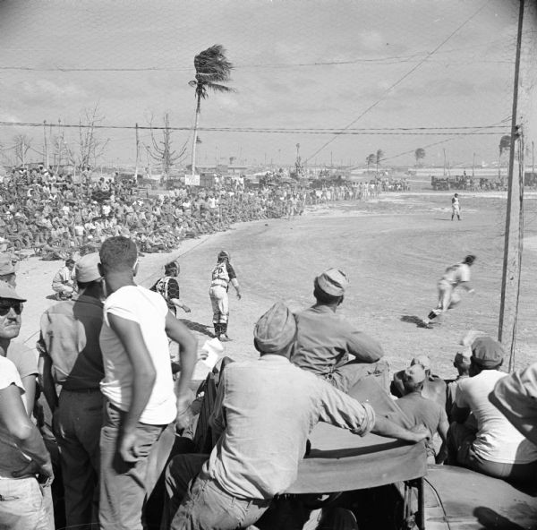 An exhibition baseball game on a Naval Base on Kwajalein Island of the Marshall Islands in the South Pacific. Both teams had Big League players drawn from navy ranks. The Navy Base declared a half holiday to allow the men to attend. The winds must have been gusty that day, as the palm fronds are streaming to the right.