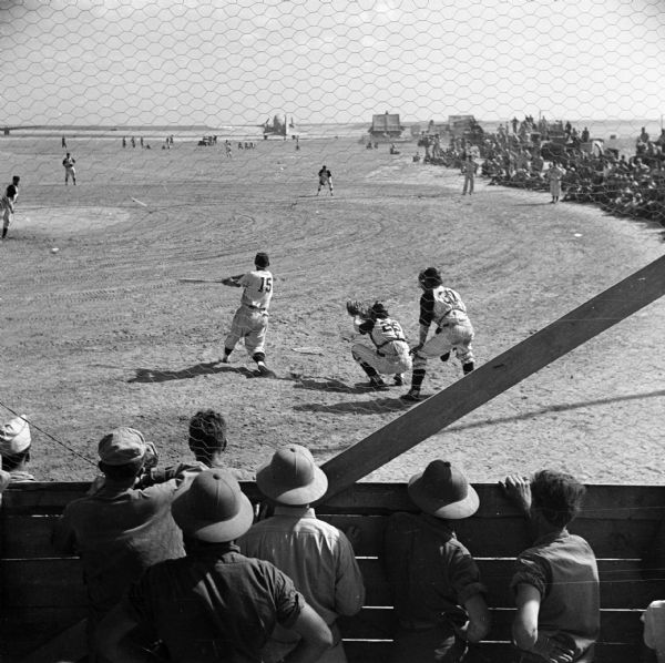 The batter swings at an exhibition baseball game on a Naval Base on Kwajalein Island of the Marshall Islands in the South Pacific. Both teams had Big League players drawn from navy ranks. The Navy Base declared a half-holiday to allow the men to attend. In the foreground spectators peer through the chicken wire fence behind home plate. On the right are more spectators, and in the distance is an airplane and several buildings near the shoreline.
