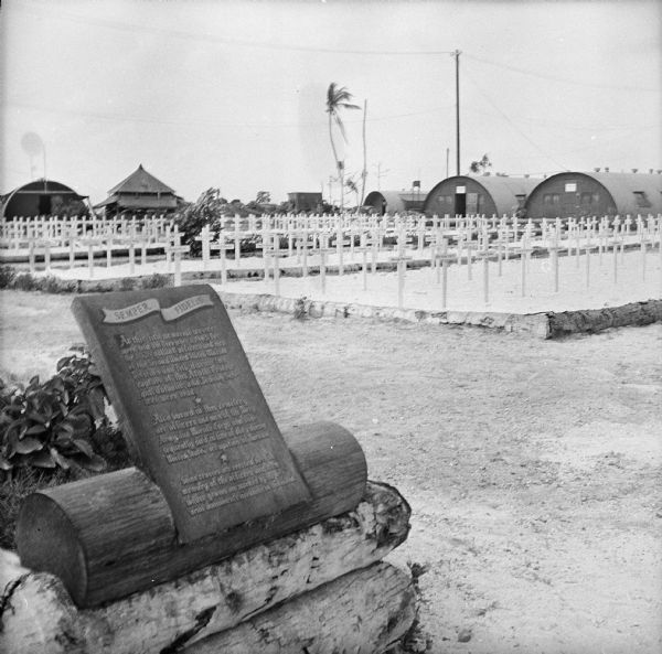 Memorial plaque and white crosses at a Marine cemetery on Tarawa Atoll in the South Pacific. Text on the plaque reads, "Semper Fidelis. At this site memorial services were held on December [1?], 1943 for the 1,026 valiant officers and men of the Second United States Marine Division who fell during the capture of this Atoll. Protestant, Catholic and Jewish Services were read. Also buried in this cemetery are officers and men of the Navy, Army and Marine Corps who subsequently died in line of duty during United States occupation of Tarawa. Some crosses are erected here in memory of the officers and men whose graves are marked by memorial monuments elsewhere on this island." Buildings are in the background.