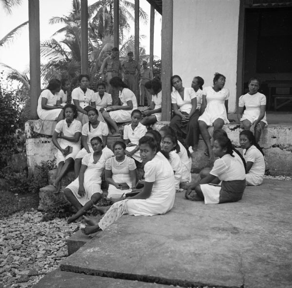 A group of students at the London Missionary Society School, Tutuila Mission located in Tutuila, American Samoa. The group is posed sitting on the concrete floor of the surrounding porch and sidewalk, and are wearing white dresses and are barefoot. Three servicemen stand on the porch on the left. Palm trees are in the background. Robert Doyle wrote a caption for this image although it was not published at that time, "Mission school for girls 14 to 22. Girls live in large dormitory. Here they study on porch. They sleep side by side on woven mats on floor."