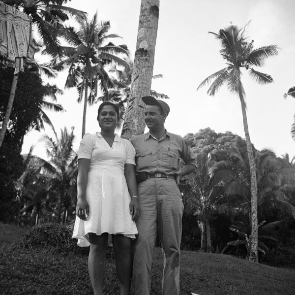 Robert Doyle poses next to a young woman from the London Missionary Society School, Tutuila Mission, located on Tutuila, American Samoa. She is wearing a white dress. A lawn and palm trees are in the background.

