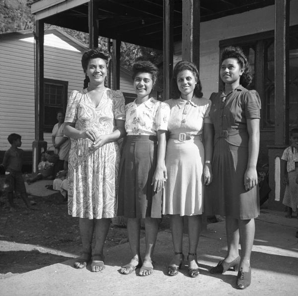 Four indigenous women pose in front of a store on Tutuila in American Samoa. They are all wearing dresses. A woman and children can be seen in the background on the left and right. Robert Doyle wrote a caption for this image although it was not published at that time, "These Samoan girls are clerks in general store at Pago Pago operated by B.F. Kneubuhl, native of Burlington, Iowa, who came to Pago Pago in 1907 and married a Samoan woman."
