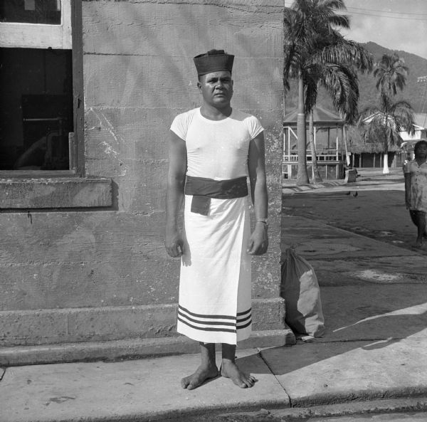 A Fita Fita Guard poses in his uniform of red cap, white skivvy shirt and white lava lava (a Samoan kilt). Shoes are not worn. The Fita Fita Guards are a special navy unit of indigenous Samoans. He is standing on the sidewalk surrounding a concrete building with an open window. A full cloth bag leans against the corner. More buildings, hills, trees and an indigenous woman are on the right. The base was located on Tutuila, American Samoa. Robert Doyle wrote a caption for this image although it was not published at that time, "Talamama, seaman, second class, is member of Fita Fita guard (U.S. navy honor guard). Stripes around bottom of lava lava denote rank. Talamama gets regular navy pay, plus 20% overseas pay, although he is not permitted to serve outside his home islands of American Samoa. Talamama was right on ball, demanded to see my credentials before he would permit me to take his picture."