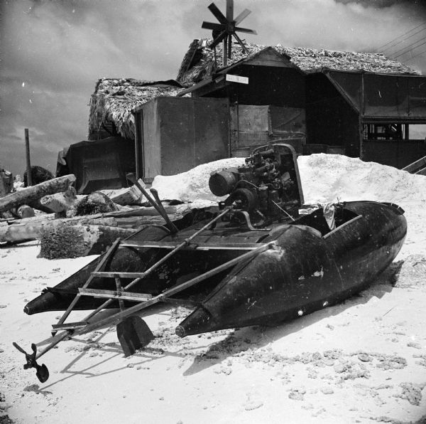 A homemade motorboat on the beach on Tarawa Atoll in the South Pacific. Robert Doyle notes that "belly tanks" were used in the boat construction. A building with a windmill is in the background.